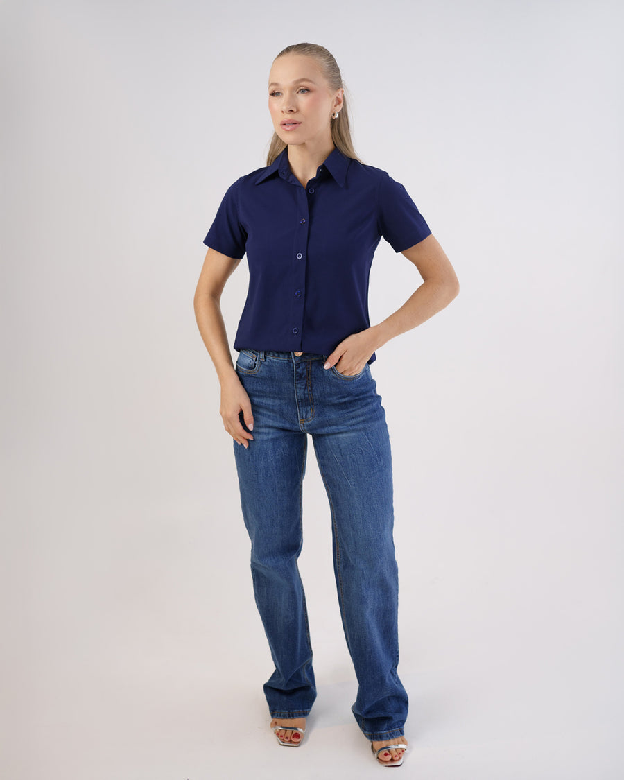 Camisa social feminina manga curta azul marinho, corpo inteiro frontal com mão no bolso da calça jeans