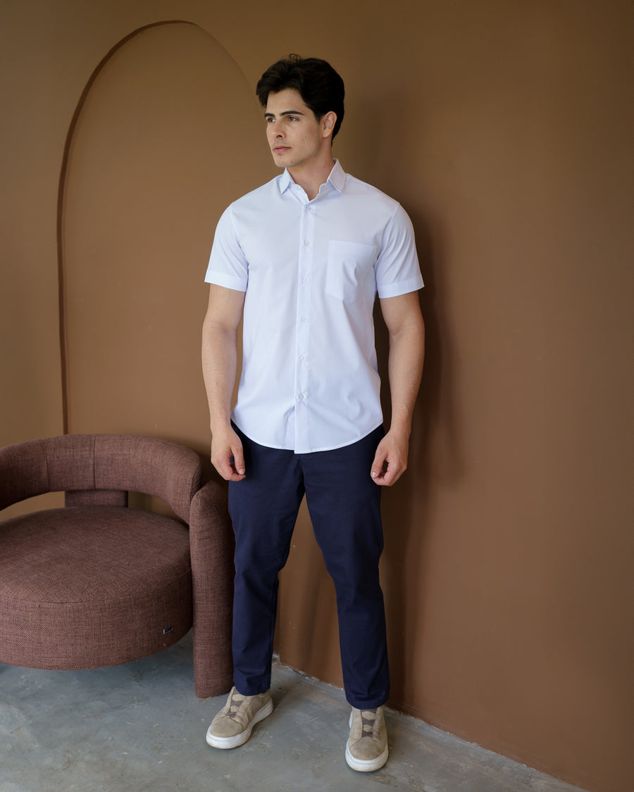Camisa social masculina manga curta branca em corpo inteiro, modelo em pe ao lado de poltrona com calca azul