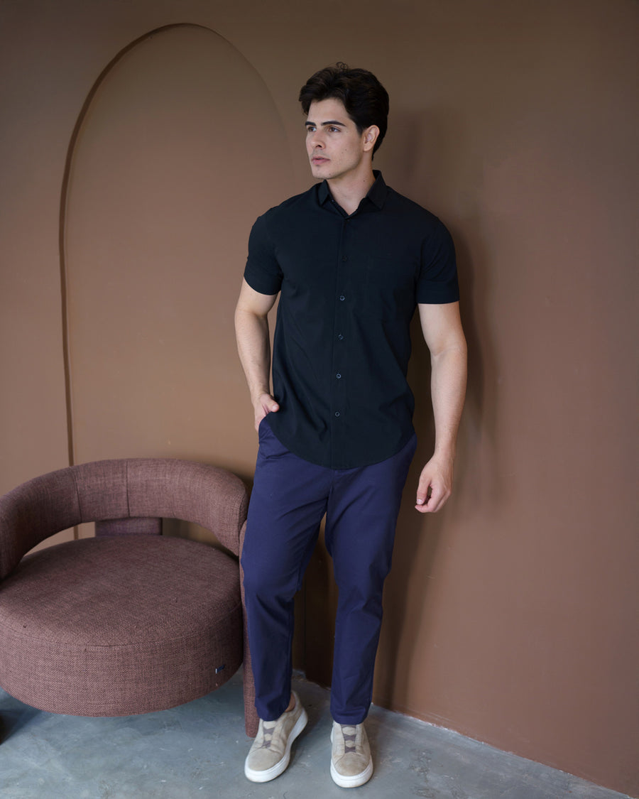 Camisa social masculina manga curta preta em corpo inteiro, mao no bolso da calca ao lado de poltrona