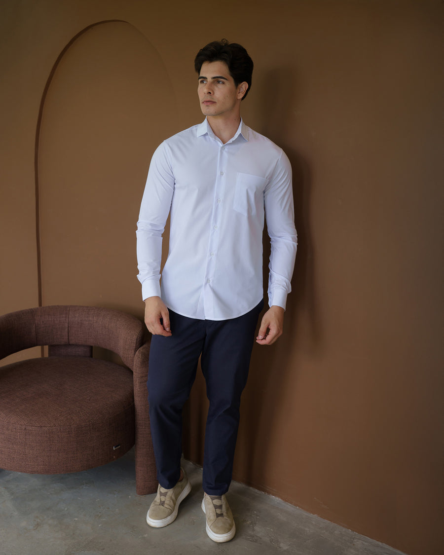 Camisa social masculina manga longa branca em corpo inteiro, modelo em pe ao lado de poltrona com calca azul