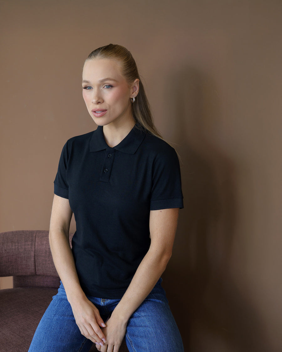 Camiseta polo feminina preta, meio corpo frontal, modelo sentada mostrando gola e botões da peça