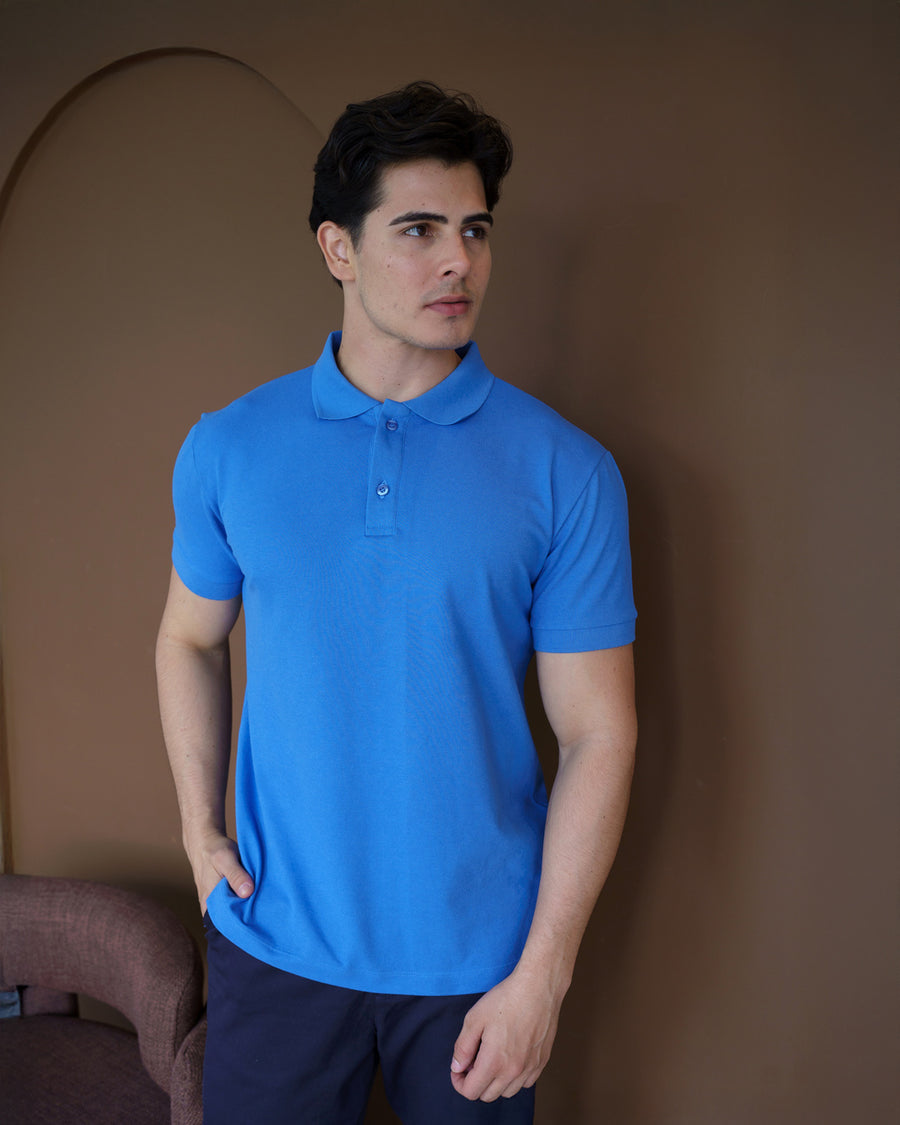 Camiseta polo masculina azul celeste em piquê, meio corpo frontal em ambiente marrom, mão no bolso