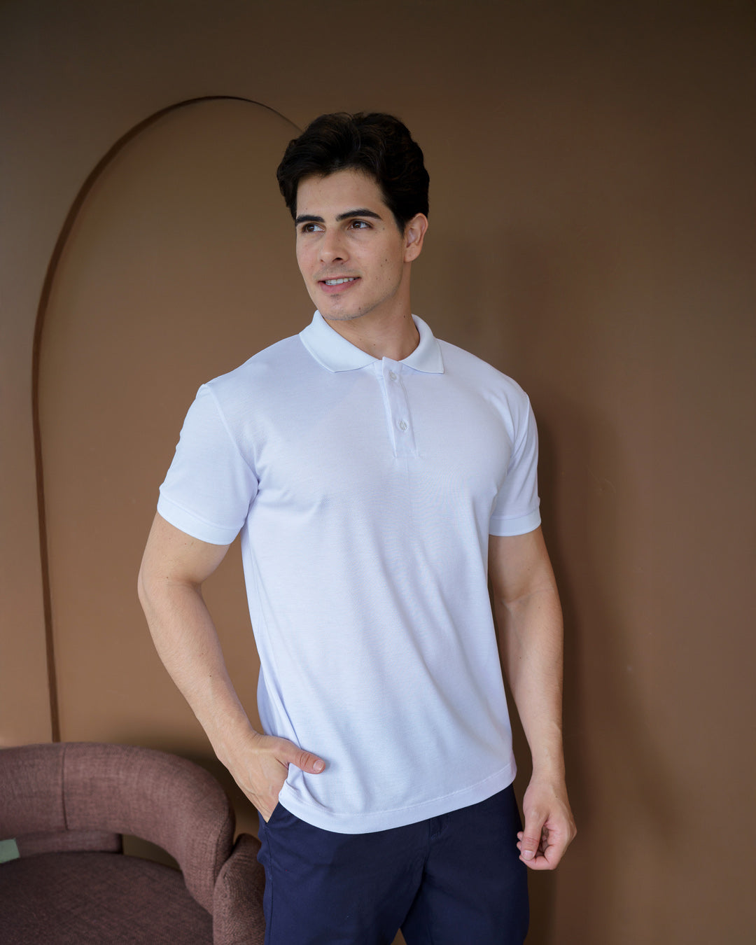 Camiseta polo masculina premium branca de frente em ambiente interno, modelo sorrindo com mão no bolso da calça — foto 2