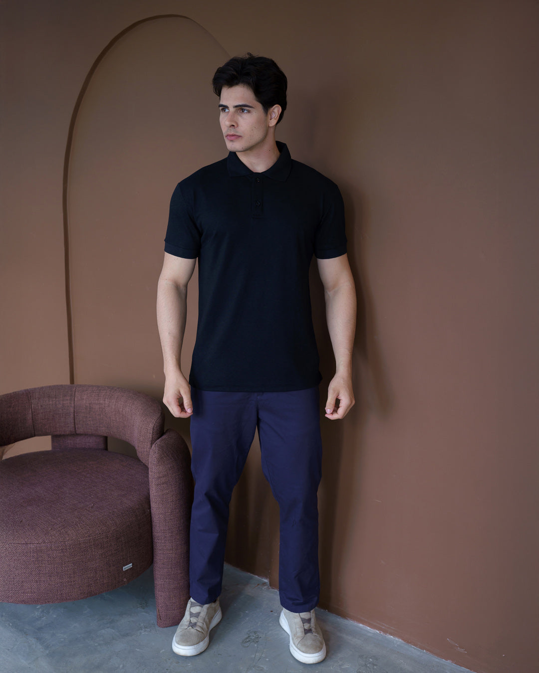 Camiseta polo masculina premium preta de corpo inteiro em cenário com poltrona, combinada com calça azul-marinho — foto 2