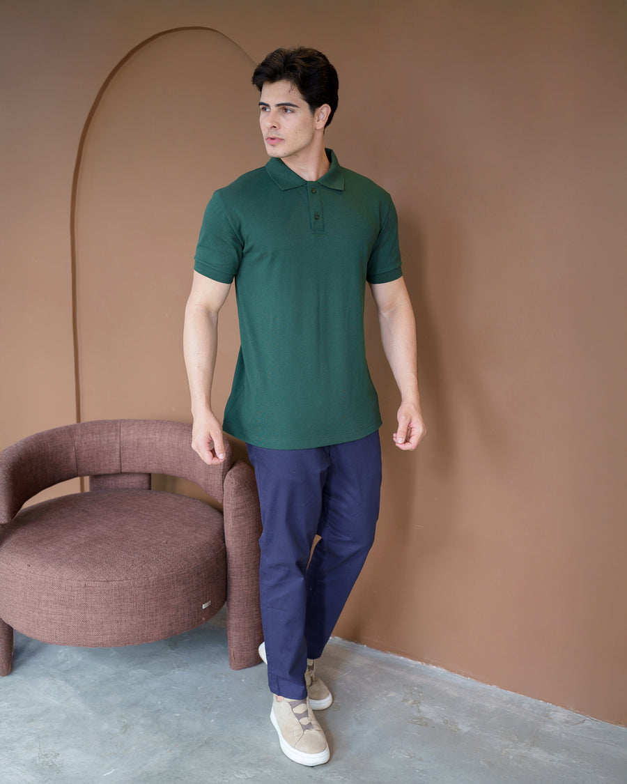 Camiseta polo masculina premium verde musgo de corpo inteiro em ambiente interno, ao lado de poltrona rosé