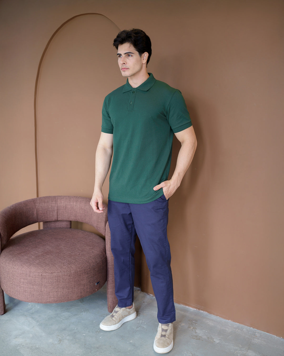 Camiseta polo masculina premium verde musgo de corpo inteiro em cenário com poltrona, modelo com mão no bolso — foto 2