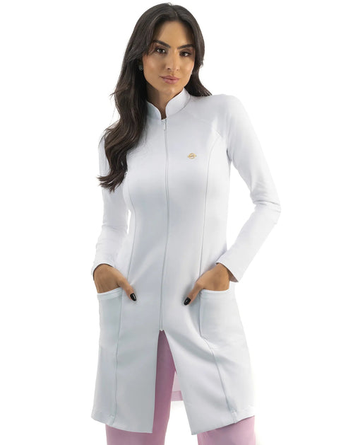 Jaleco feminino Vicky branco de frente, gola alta com zíper e pin dourado, modelo com as mãos nos bolsos frontais