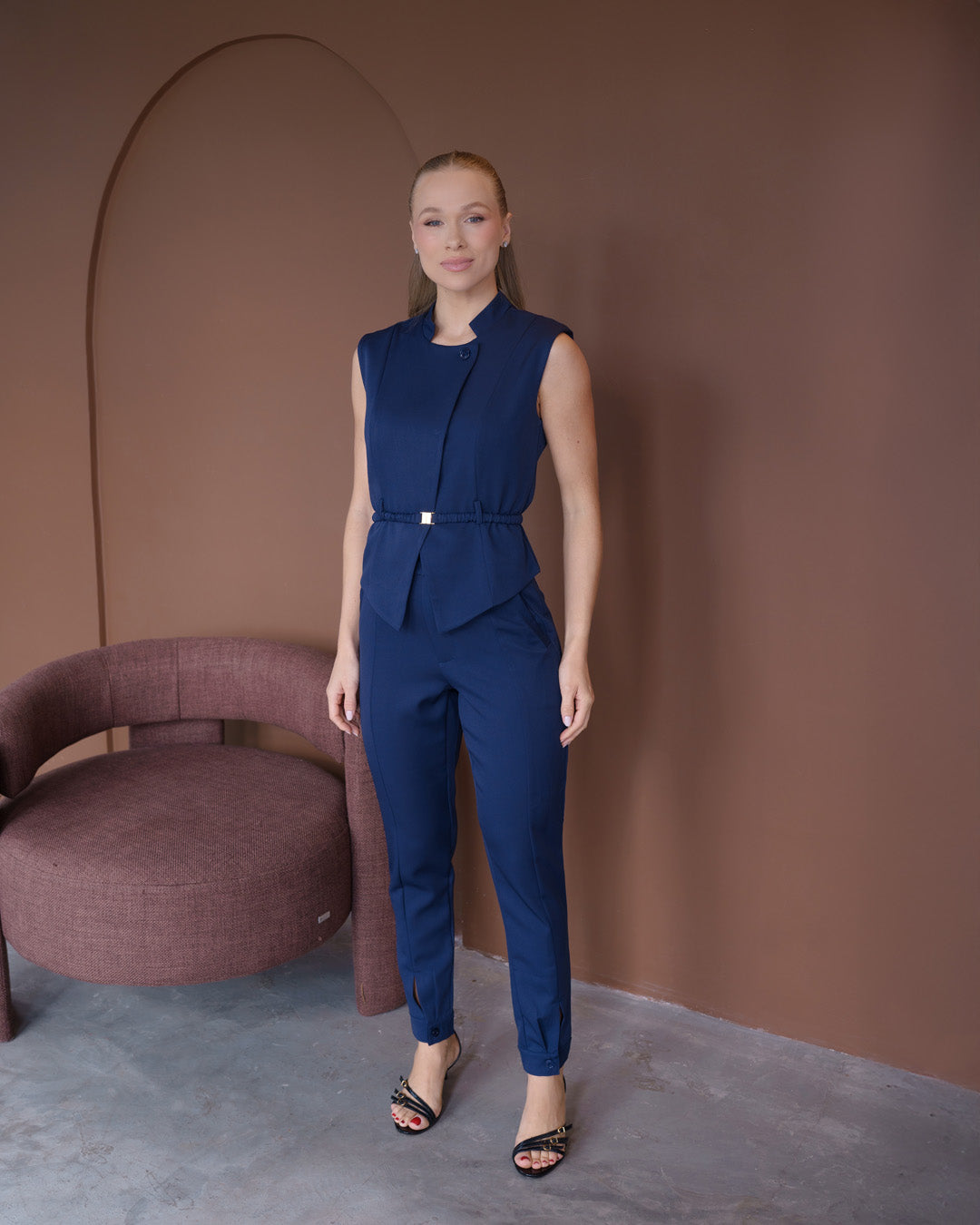 Scrub feminino Ella azul marinho de frente em cenário com poltrona, blusa com cinto de fivela dourada e calça slim — foto 3