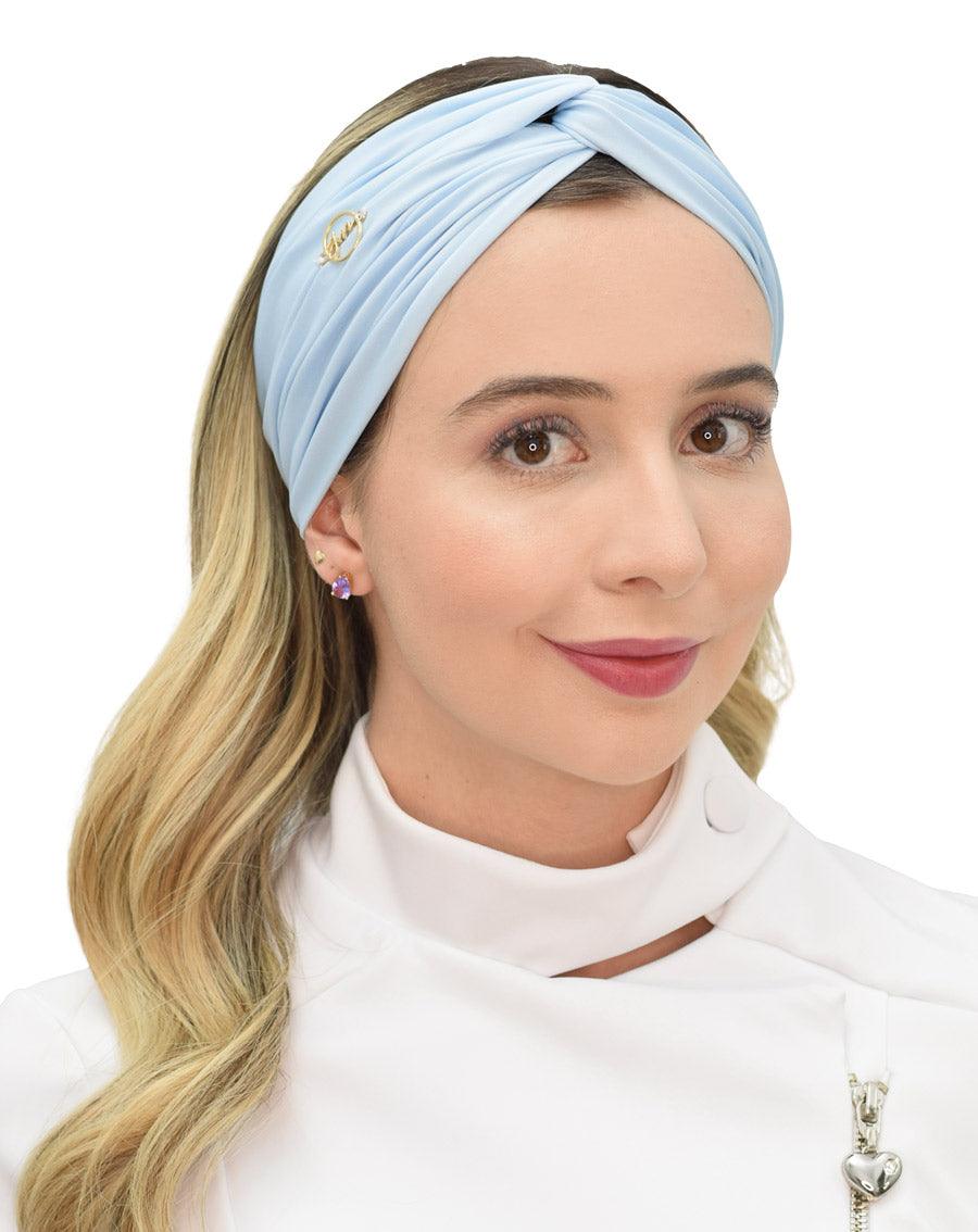 Turbante feminino azul claro em modelo, vista frontal, com franzido cruzado na frente e pin dourado da marca