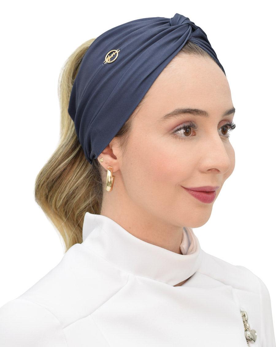 Turbante feminino azul marinho em modelo, vista lateral, com nó torcido no alto e pin dourado da marca