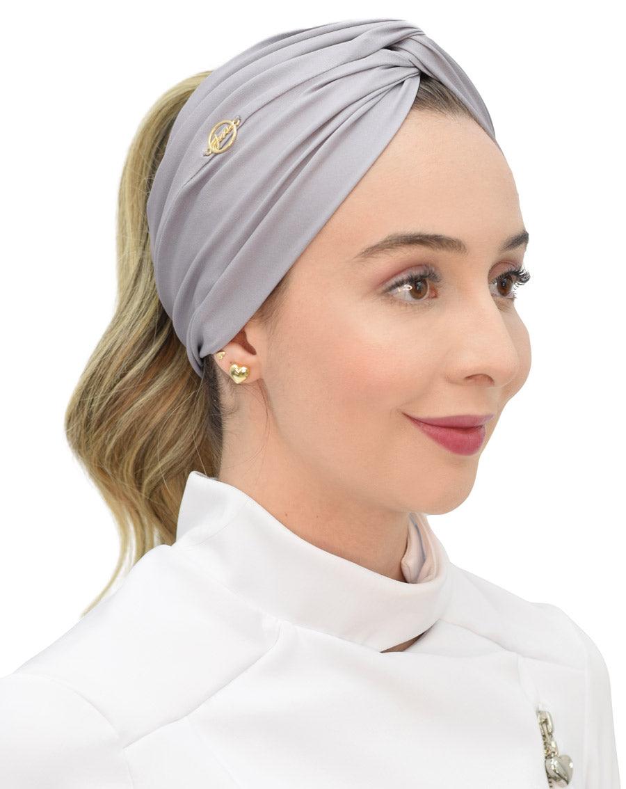 Turbante feminino cinza claro em modelo, vista lateral, tecido acetinado com nó cruzado e pin dourado da marca