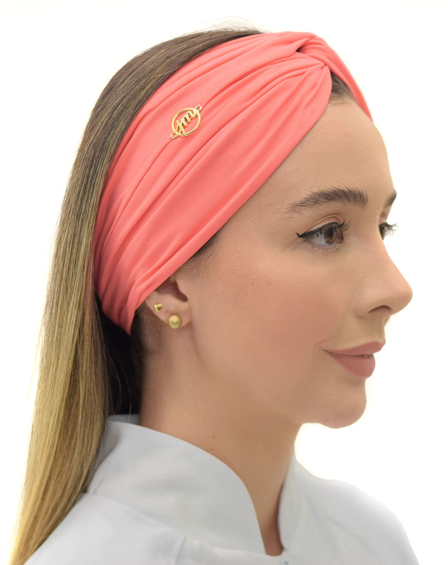 Turbante feminino goiaba em modelo de perfil, tecido coral drapeado cobrindo a cabeça e pin dourado da marca