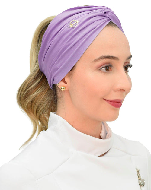 Turbante feminino lavanda em modelo de perfil, tecido lilás drapeado, logo dourado bordado e cabelo em rabo