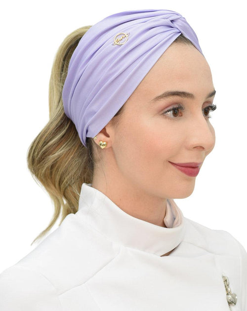 Turbante feminino lilás claro em modelo de perfil, tecido drapeado cobrindo a cabeça e logo dourado bordado