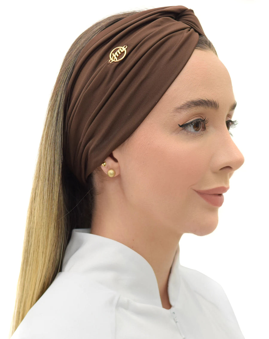 Turbante feminino marrom em modelo de perfil com cabelo solto, tecido franzido cobrindo a cabeça e pin dourado