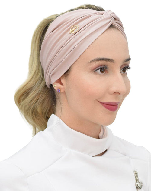 Turbante feminino nude em modelo, vista lateral, tecido rosé drapeado com nó no alto e pin dourado da marca