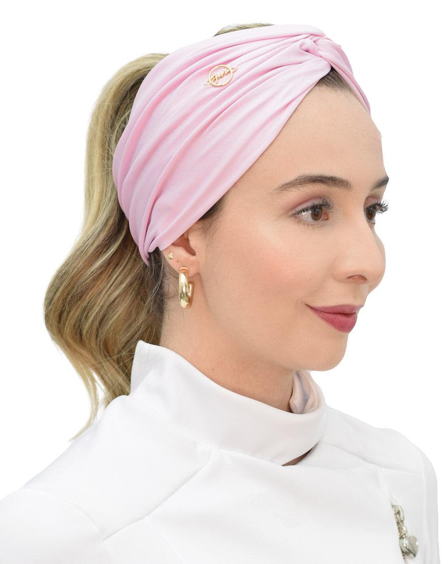 Turbante feminino rosa claro em modelo de perfil, tecido drapeado cobrindo a cabeça e logo dourado da marca