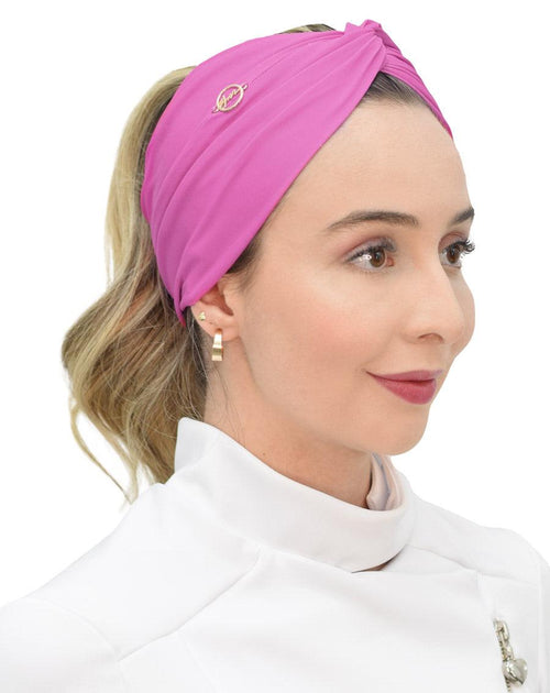 Turbante feminino rosa fúcsia em modelo, vista lateral, tecido drapeado com nó no alto e pin dourado da marca