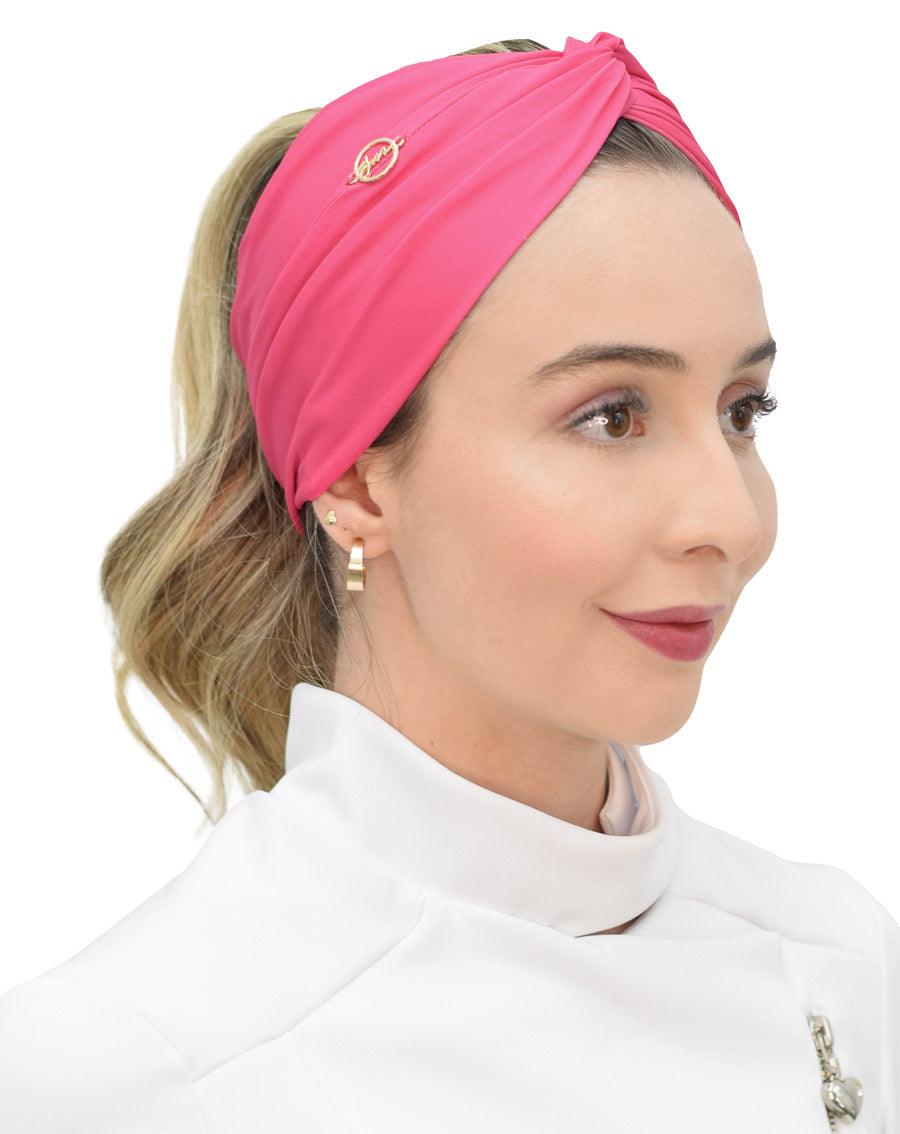 Turbante feminino rosa pink em modelo, vista lateral, tecido drapeado com nó no alto e pin dourado da marca