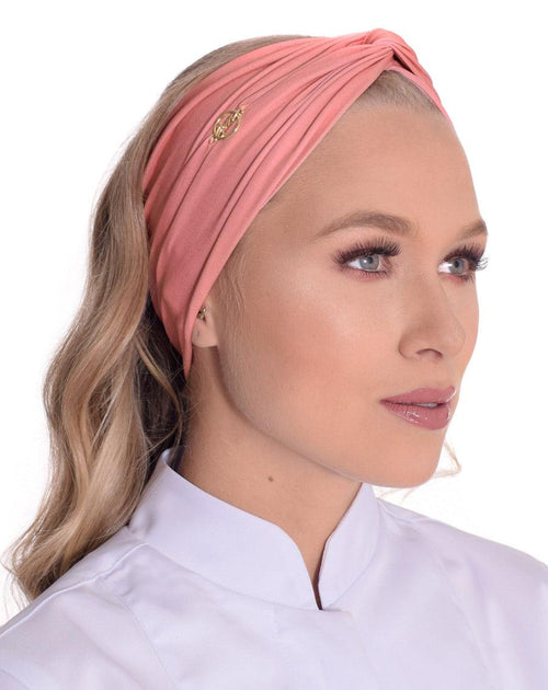 Modelo usando turbante feminino rosé de malha franzida com nó frontal e emblema dourado, sobre jaleco branco