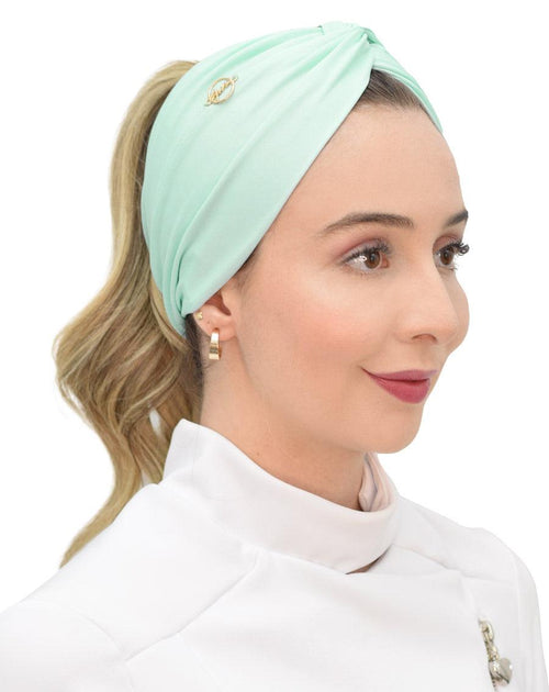 Modelo de rabo de cavalo usando turbante feminino verde água com nó frontal e emblema dourado, sobre jaleco branco