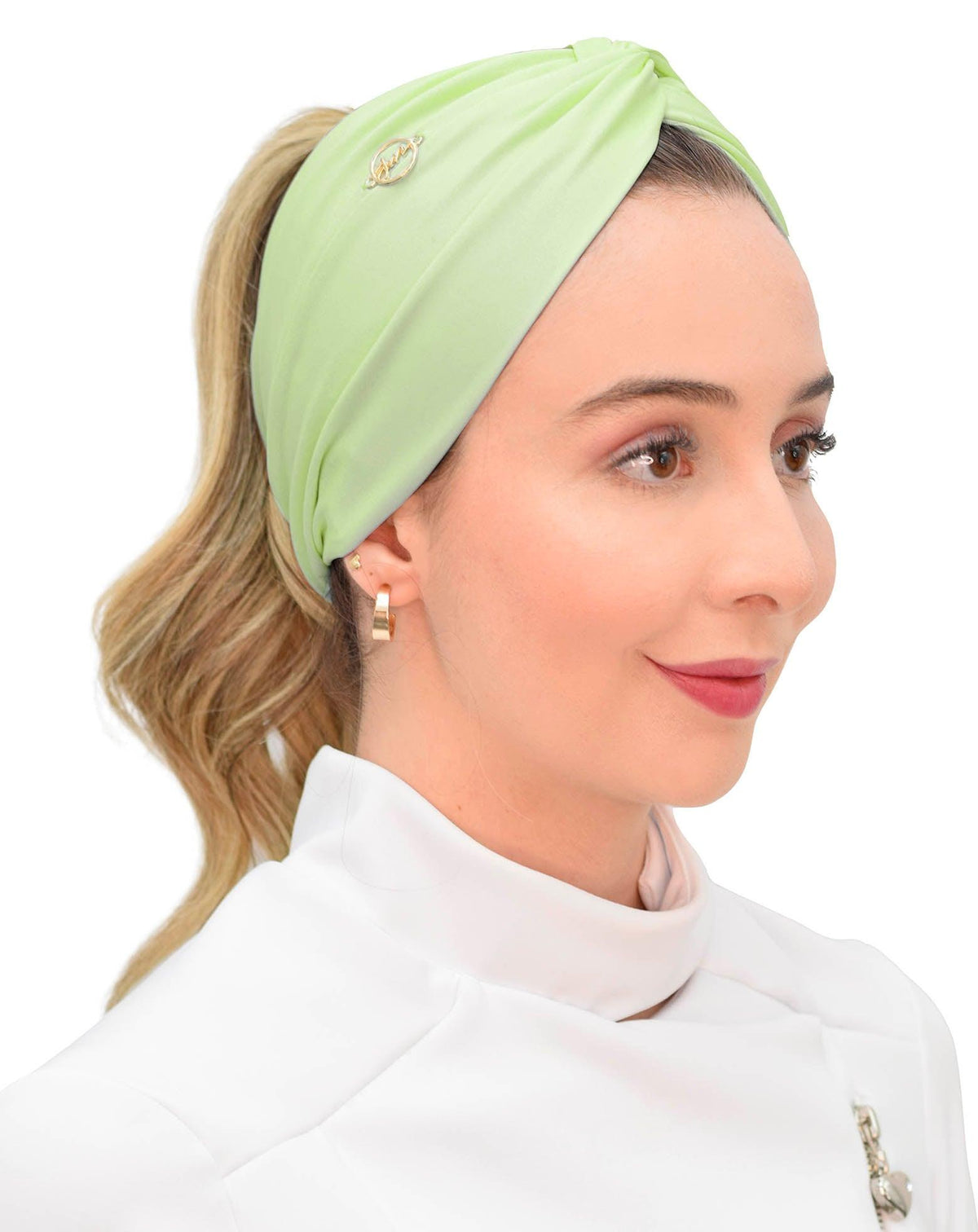 Modelo de rabo de cavalo usando turbante feminino verde pistache com nó frontal e emblema dourado, sobre jaleco branco